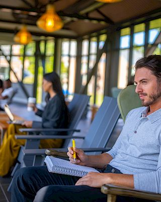 A man sits in a chair holding a notebook and pen, while others are seated in the background using laptops and chatting.