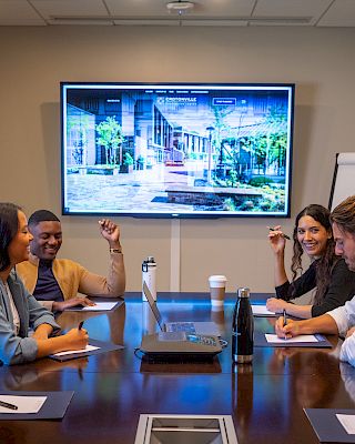 A group of four people are in a conference room, sitting around a table with notebooks, a screen, and a flip chart.