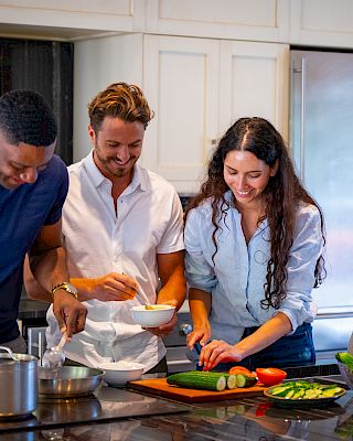 Four people are happily cooking together in a modern kitchen, chopping vegetables and preparing food near the stove.