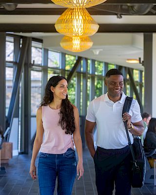 Two people are walking and smiling in a modern hallway with large windows and pendant lights. Others are seated, engaging with devices.