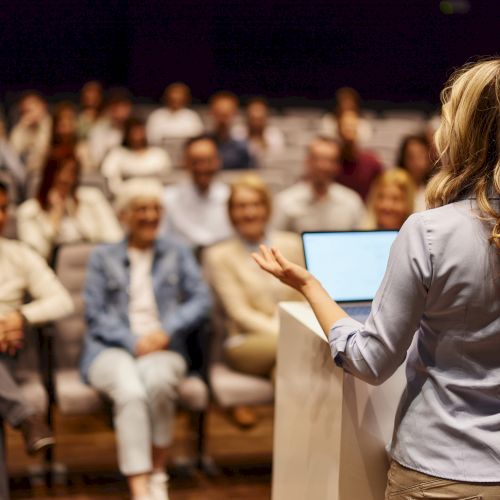 A woman is speaking at a podium to an audience seated in rows, suggesting a seminar or presentation setting.