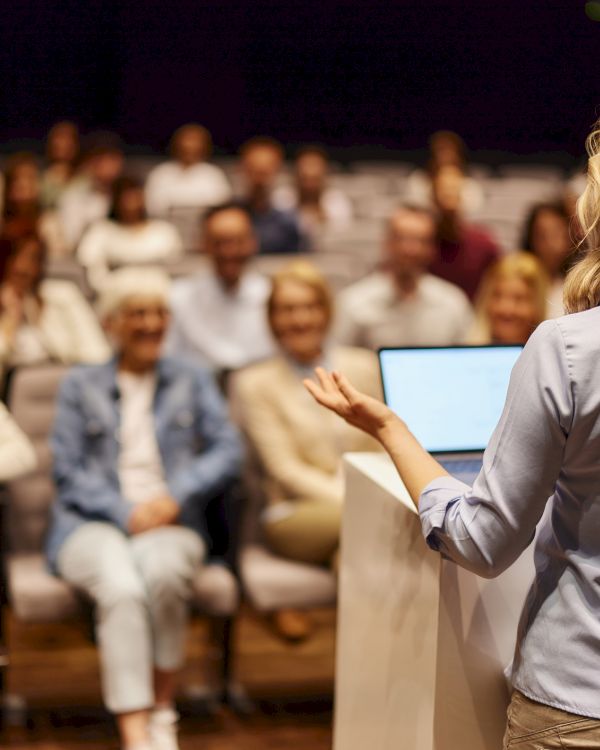 A woman is speaking at a podium to an audience seated in rows, suggesting a seminar or presentation setting.