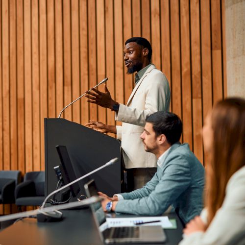 A person is speaking at a podium in a conference room with a panel of seated individuals on the side, focused on the presentation.