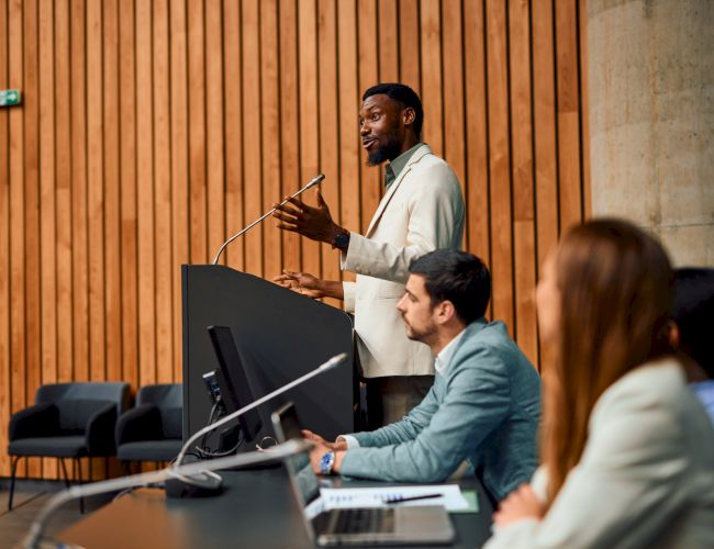 A person is speaking at a podium in a conference room with a panel of seated individuals on the side, focused on the presentation.