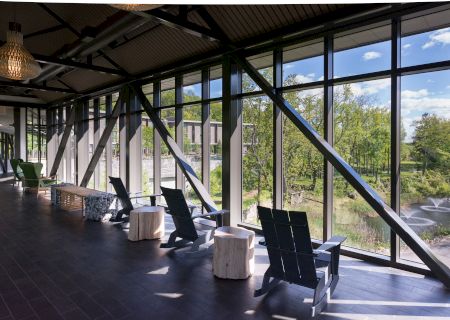 A sunlit indoor seating area with large windows overlooks a scenic outdoor view with greenery and a pond with fountains.