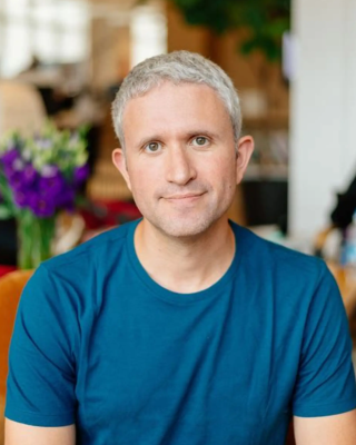 A person with short gray hair in a blue shirt sits in a room with blurred background and flowers.