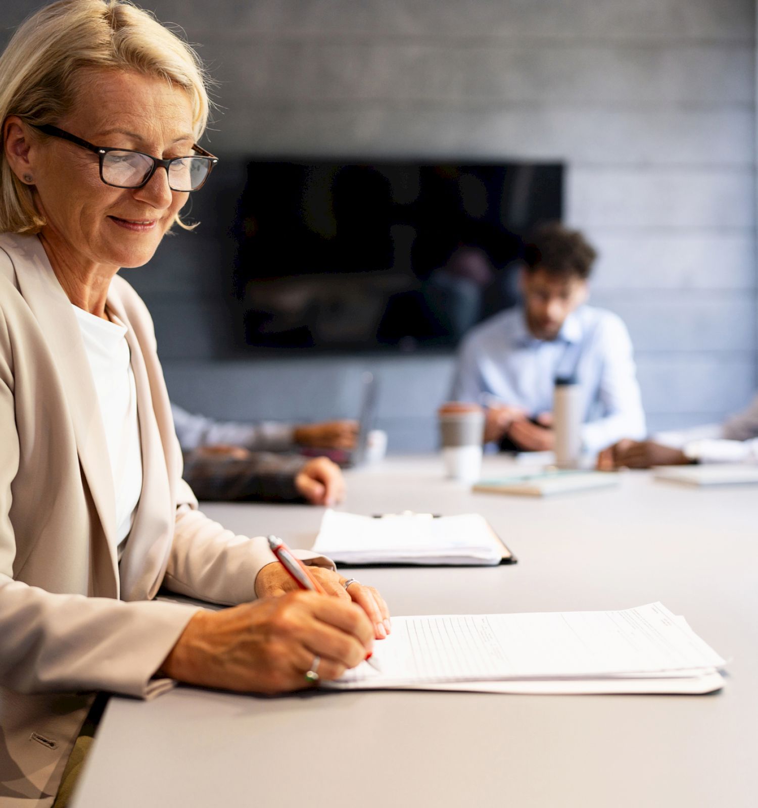 A woman in glasses is writing at a conference table, while three people in the background are engaged in discussion.